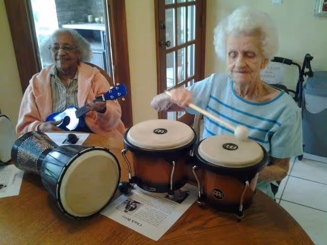 Two elderly women sitting at a wooden table playing musical instruments. One woman is playing a small blue guitar or ukulele, and the other is playing a pair of bongos with a mallet. There is also a drum on the table. The setting appears to be indoors with a door and window in the background.