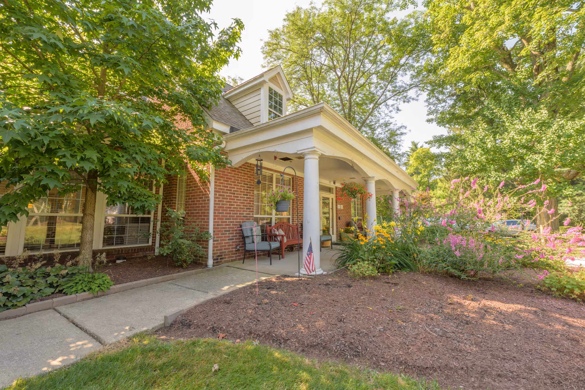 Front porch area of a brick building with white columns, outdoor chairs, hanging flower pots, and a garden with various plants and flowers. Trees surround the area, and a small American flag is planted in the garden bed.
