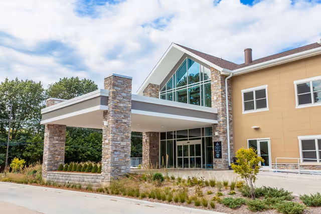 Exterior view of Cottage Grove Place, showing a modern building entrance with stone pillars supporting a covered drop-off area. The building has large windows and beige siding, surrounded by landscaped greenery and trees in the background.