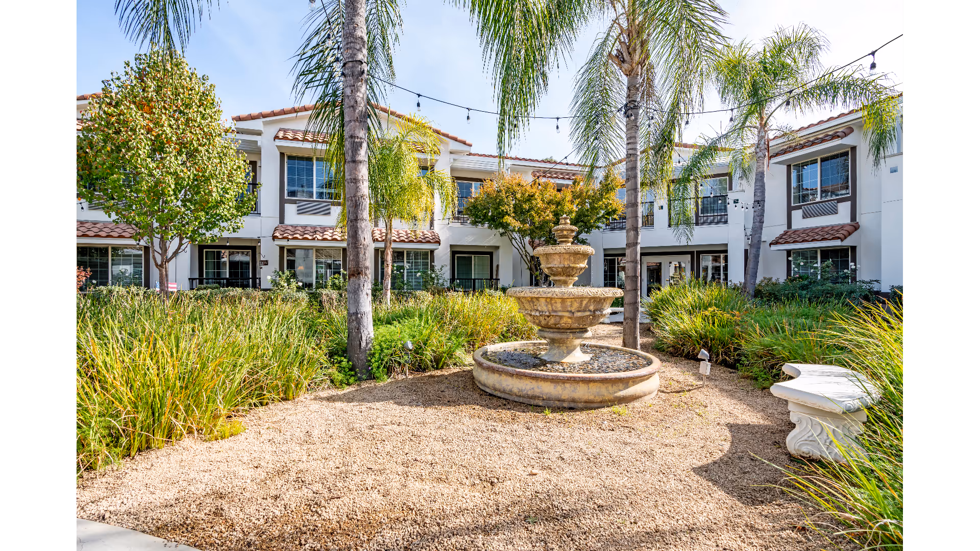 Outdoor courtyard area at Westmont of Fresno featuring a central tiered stone fountain surrounded by gravel, palm trees, green shrubs, and a white stone bench. The background shows a two-story building with white walls, brown trim, and windows with blinds.