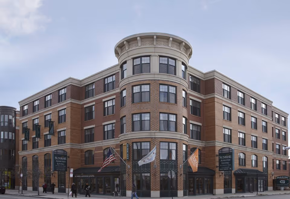Front exterior of a multi-story brick Sunrise of Lincoln Park assisted living building with flags at the main entrance.