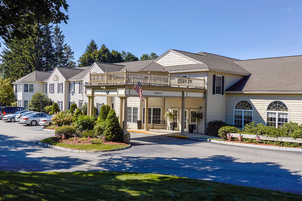 Exterior view of Benchmark Senior Living at Leominster Crossings showing the main entrance with a covered drop-off area, American flags, parked cars, and surrounding greenery under a clear blue sky.
