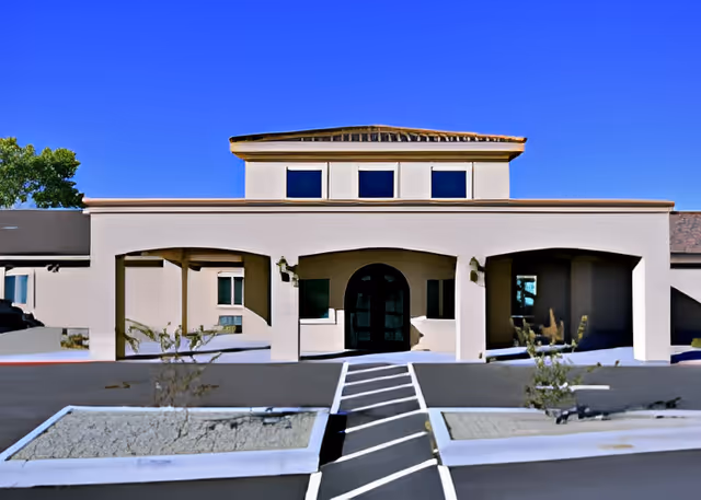 Front exterior view of a single-story assisted living facility building with a beige facade, arched entryways, and a tiled roof under a clear blue sky. There is a paved driveway and landscaped areas with small plants in front of the entrance.
