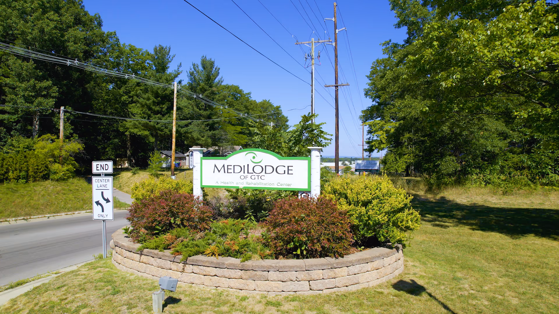 Outdoor view of the Medilodge of GTC sign surrounded by greenery and bushes, located next to a road with traffic signs and utility poles under a clear blue sky.