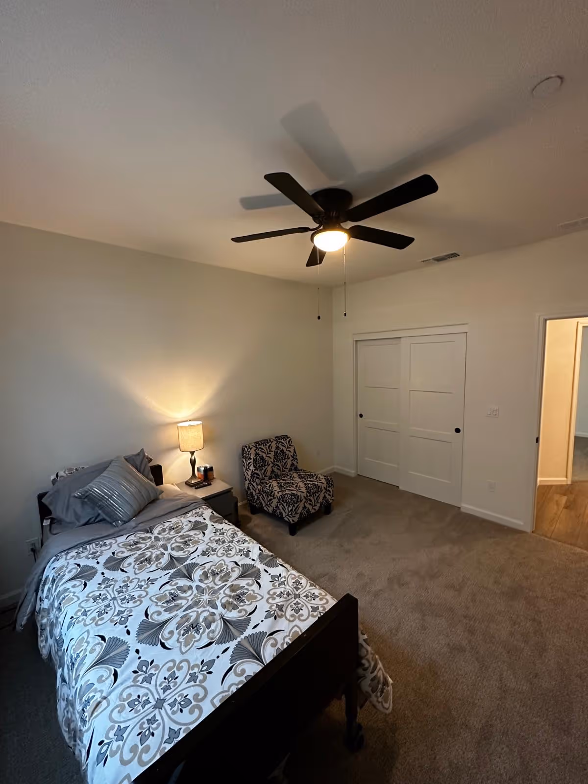 Carpeted single bedroom with a patterned bed, nightstand and lamp, an upholstered chair, ceiling fan, and double closet doors.