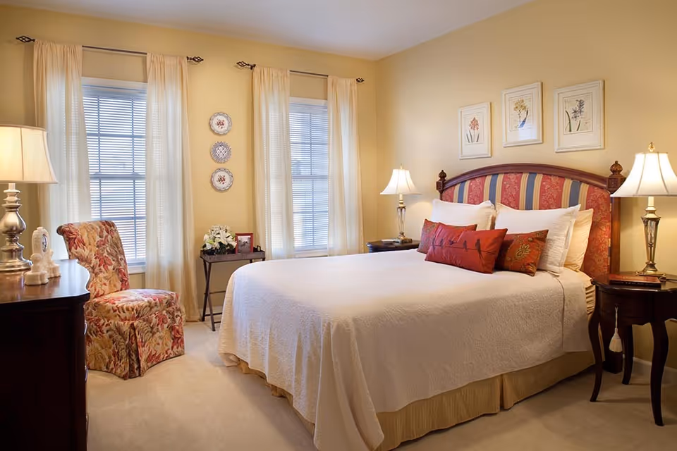 Bright traditionally furnished bedroom with a bed in white linens, striped upholstered headboard, bedside tables with lamps, two tall windows with sheer curtains, and a floral armchair.