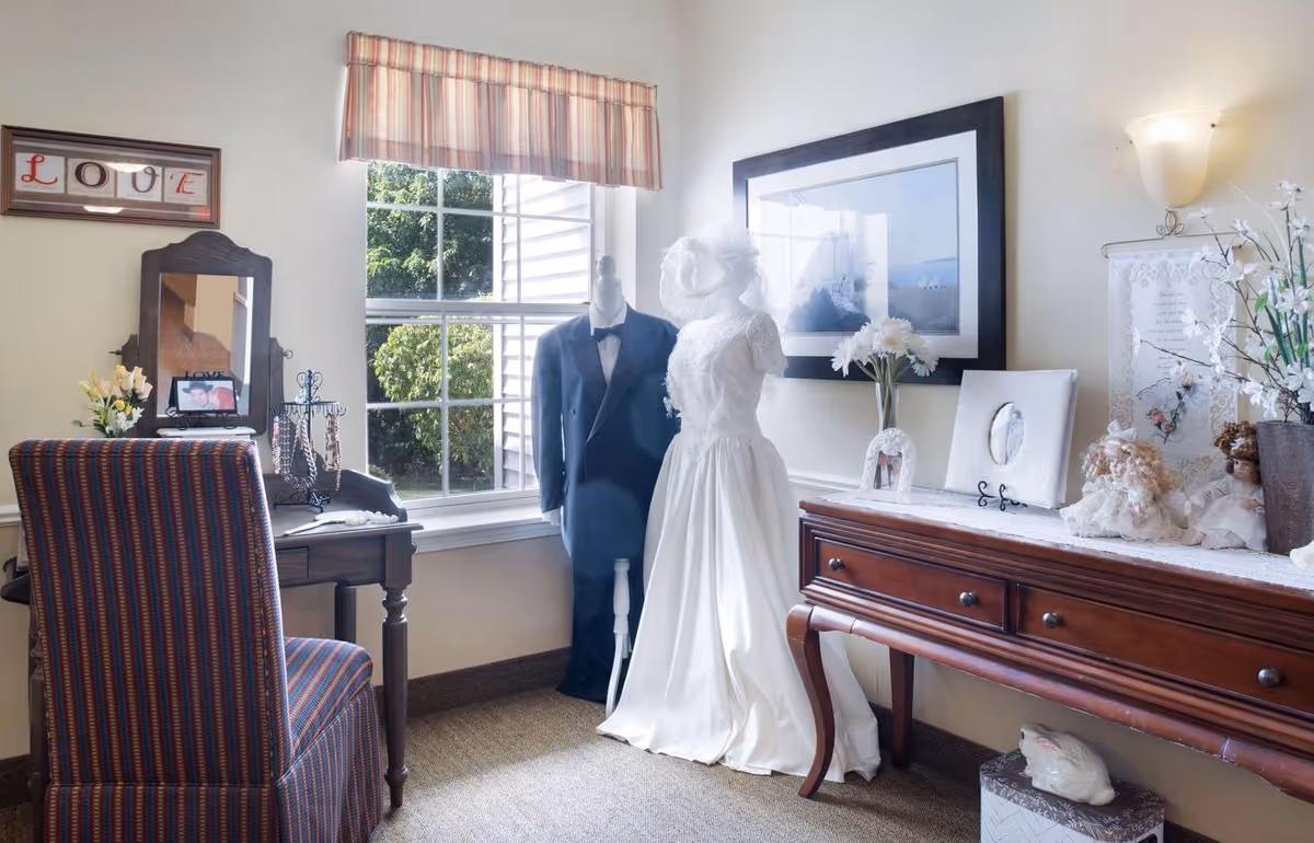 A cozy room with a window showing greenery outside. Inside, there is a striped upholstered chair in front of a wooden vanity table with a mirror and jewelry stand. Next to the window, two mannequins display a black tuxedo and a white wedding dress. A wooden sideboard holds framed photos, flowers, and decorative dolls. The walls are light-colored with a framed picture and a wall sconce light.