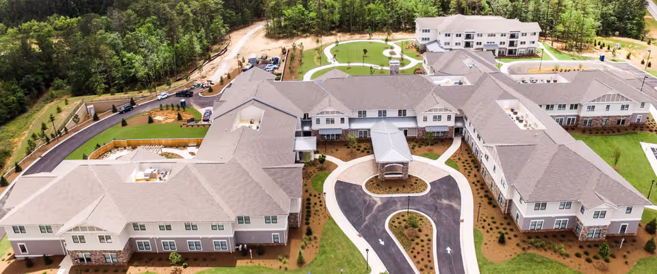 Aerial view of The Glen at Lake Oconee Village senior living facility showing a large, multi-wing building surrounded by greenery and trees. The building has multiple entrances, a circular driveway, and landscaped grounds with pathways and parking areas.