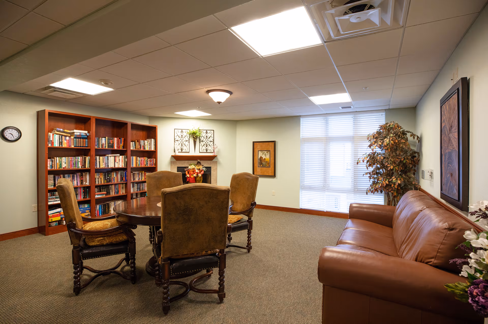 A cozy common room with a round wooden table surrounded by four upholstered chairs, a brown leather sofa, a large bookshelf filled with books, a decorative fireplace with autumn-themed decorations, a framed picture on the wall, a large window with blinds, and a potted plant in the corner.