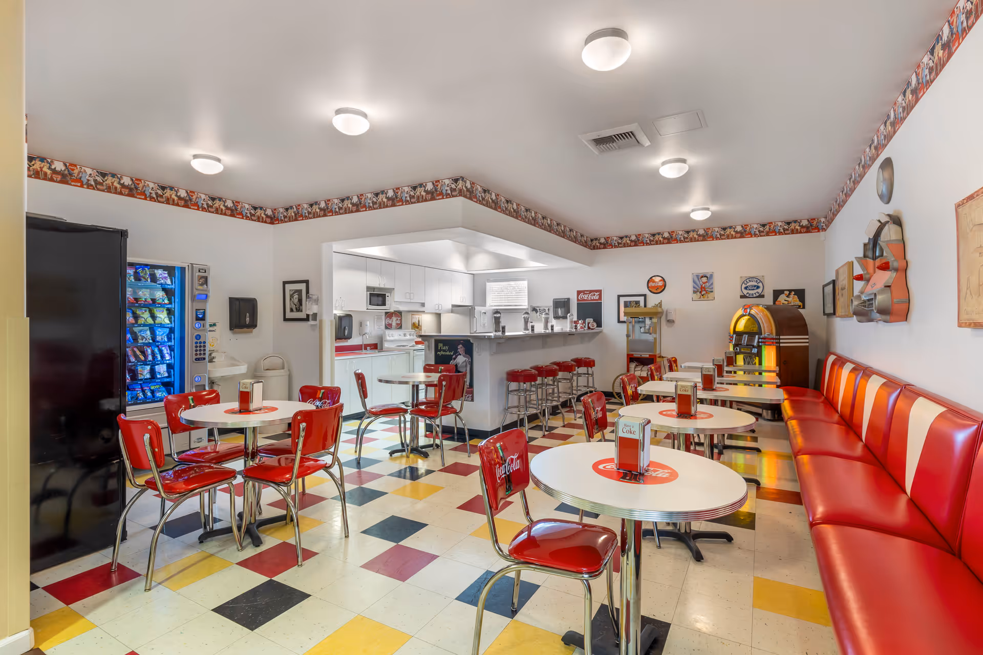 Retro-style dining room with red booths and chairs around round tables, a jukebox, vending machine, and a kitchen counter area.