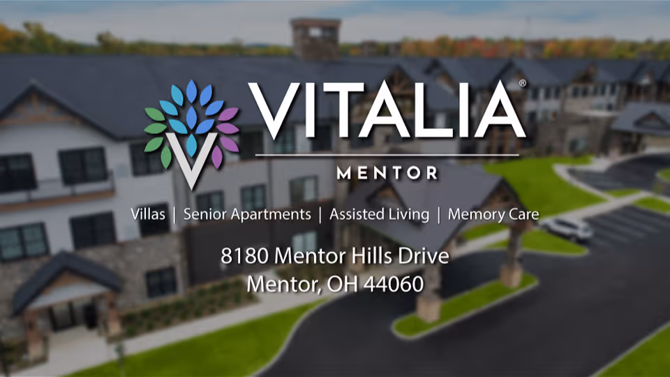Exterior view of the VITALIA Mentor senior living facility showing a multi-story building with stone and siding facade, surrounded by green lawns and paved driveways.