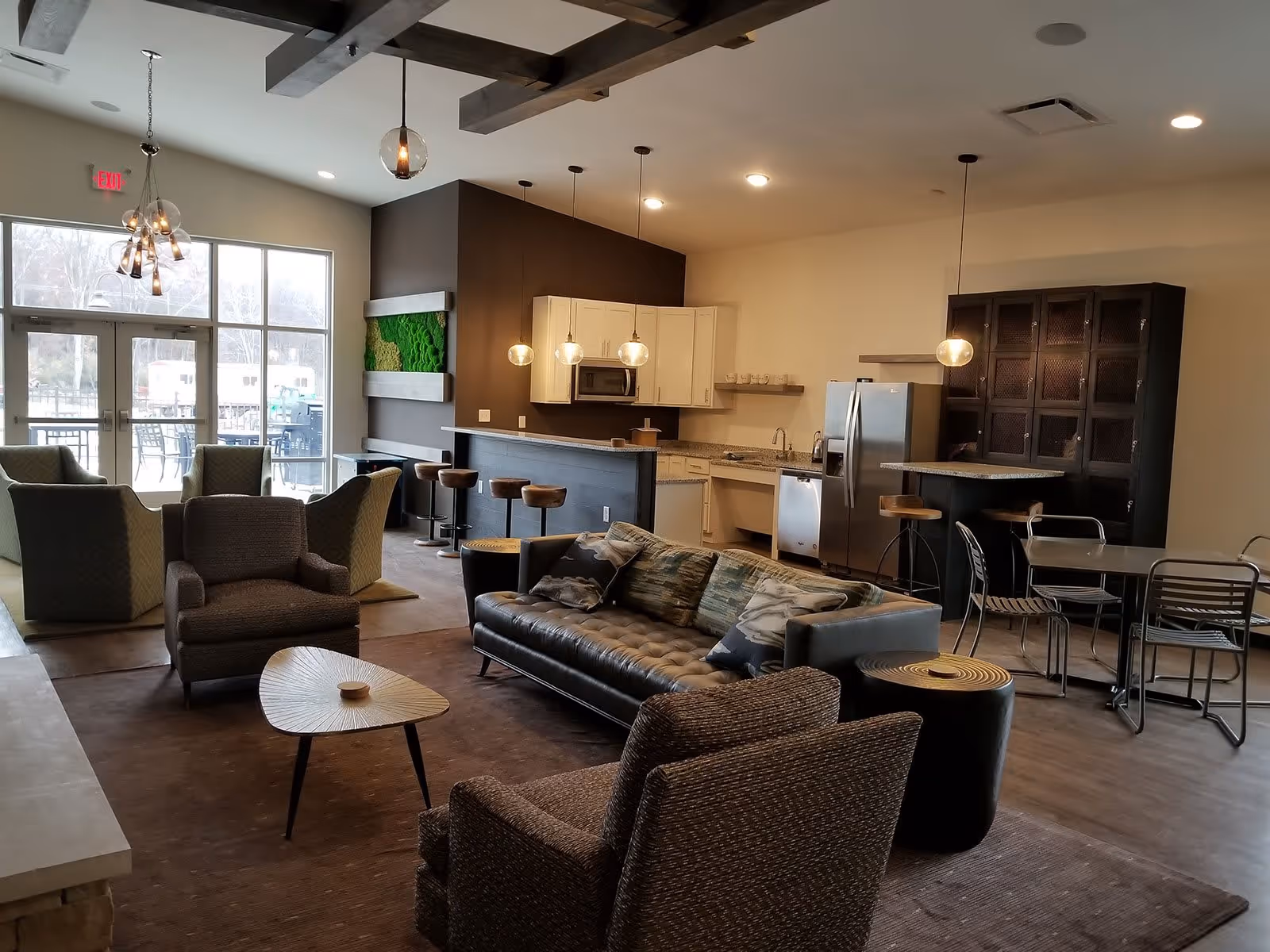 A modern senior living common area featuring a seating arrangement with armchairs and a sofa around a small coffee table. The space includes a kitchen area with white cabinets, stainless steel appliances, a bar counter with stools, and a dining table with chairs. Large windows and glass doors provide natural light and a view of an outdoor patio with tables and chairs.