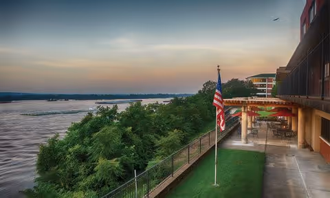 Riverside terrace at sunset with an American flag, railing, trees and a building with outdoor seating.