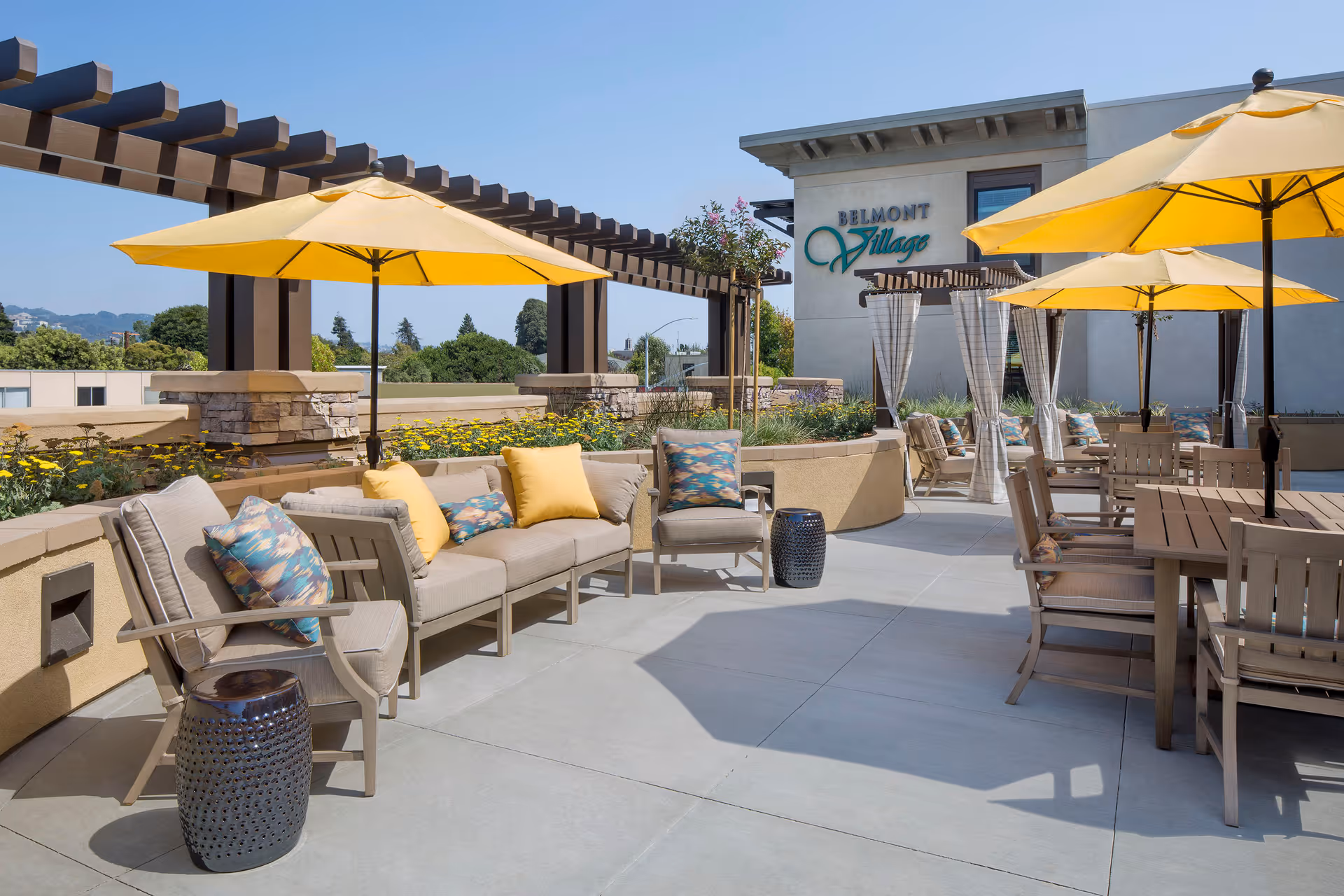 Outdoor patio area at Belmont Village Senior Living Albany featuring beige cushioned seating with colorful pillows, yellow umbrellas providing shade, wooden tables and chairs, and a pergola structure with greenery and flowers in the background under a clear blue sky.