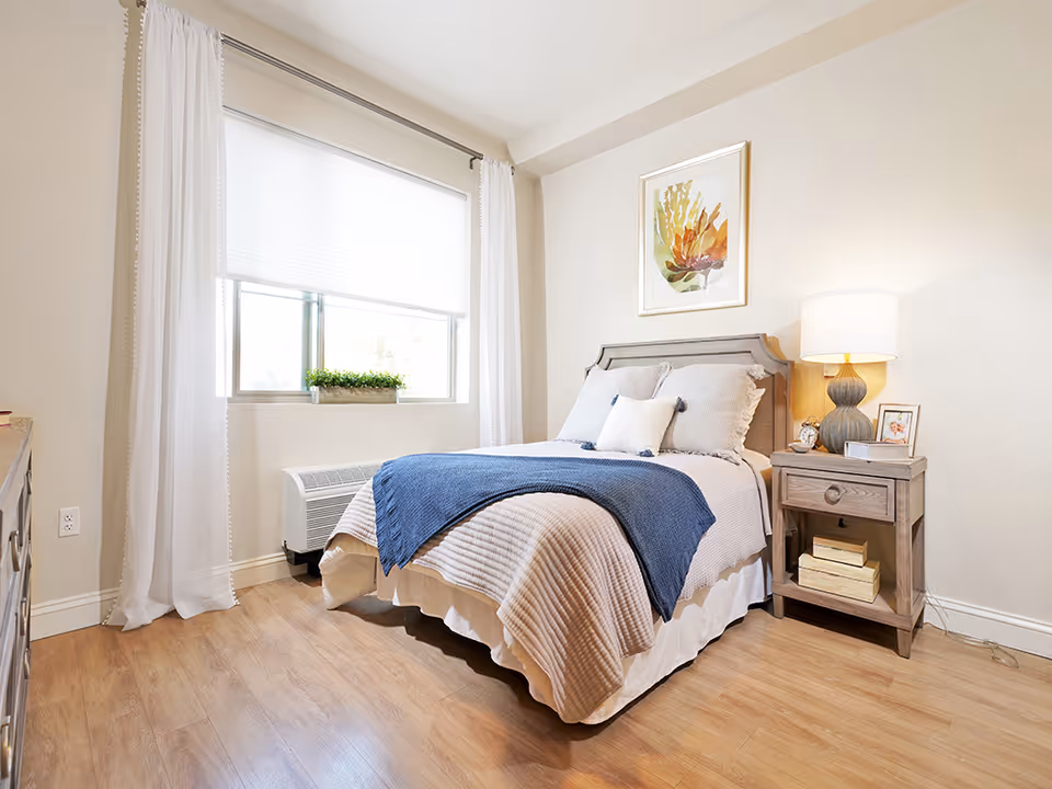 Bright, neatly furnished bedroom with a made bed, nightstand and lamp beside a window with white curtains and a small plant.