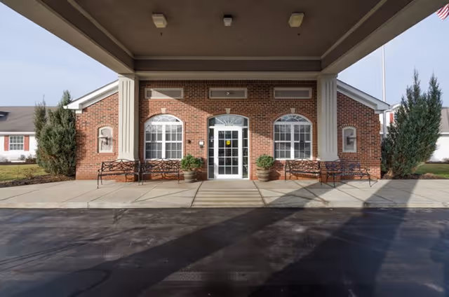 Front entrance of a brick building with a covered driveway, two large columns, two benches, potted plants, and an American flag visible on the right side.
