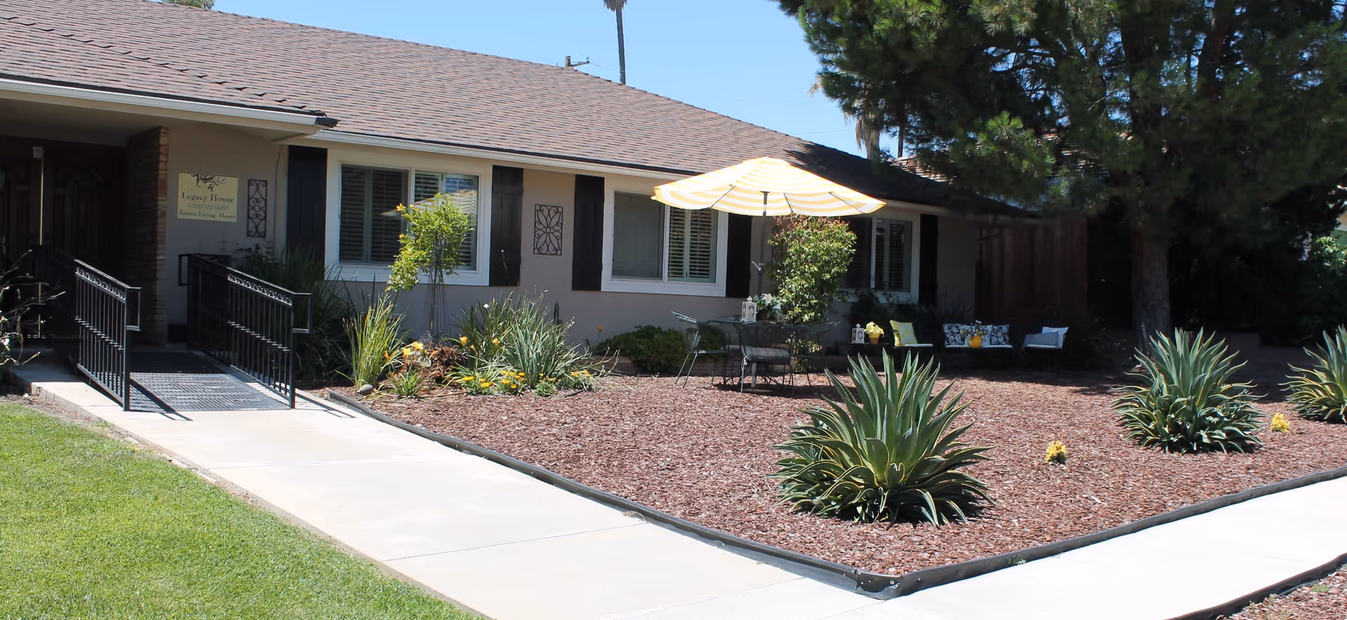 Exterior view of Legacy House Upland, a single-story building with a brown shingled roof and beige walls. The front yard features a landscaped area with mulch, several agave plants, and a patio set with a table, chairs, and a large beige umbrella. There is a concrete walkway leading to a ramp with black railings at the entrance. Trees and shrubs surround the building.