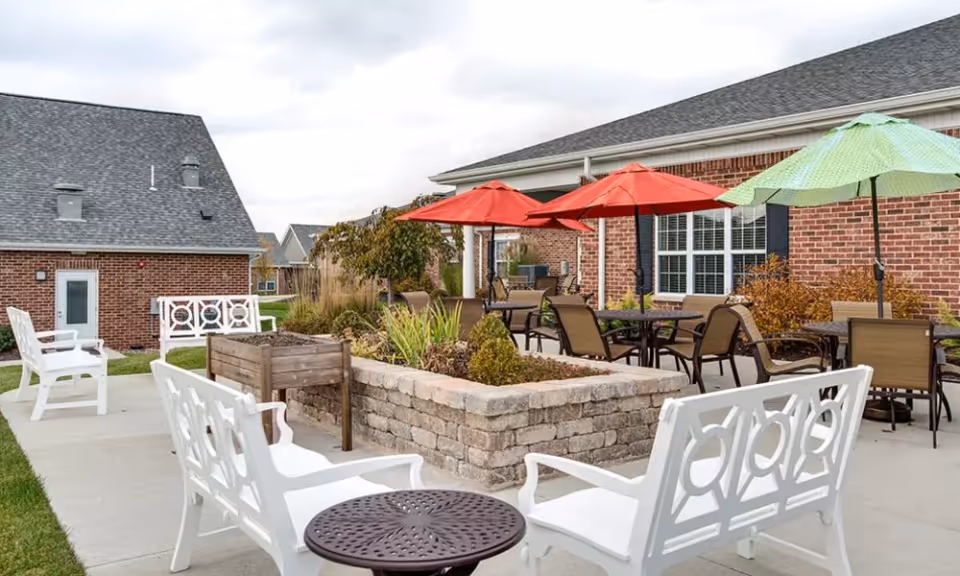 Outdoor patio area at Mill Creek Village Senior Living featuring white benches, round tables with chairs, and colorful umbrellas in red and green. The patio is surrounded by a low stone planter with greenery, and brick buildings are visible in the background under a cloudy sky.