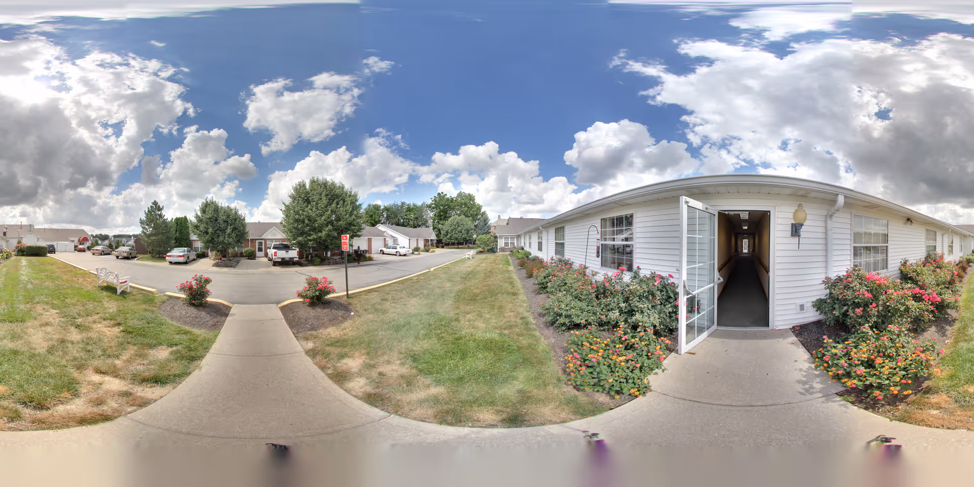 Outdoor view of a senior living facility with a concrete walkway leading to an open door of a single-story white building. The building is surrounded by well-maintained flower beds with colorful flowers and bushes. Across the street, there are parked cars and more single-story buildings with trees and a partly cloudy sky above.