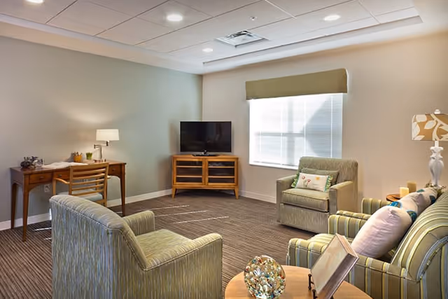 A bright communal living room with upholstered chairs and a sofa around a coffee table, a TV on a wooden stand, a desk with a lamp, and a large window.