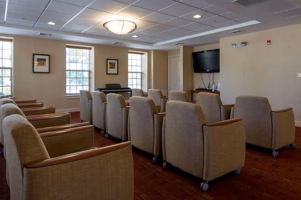 A room with two rows of beige upholstered chairs with wooden armrests, arranged facing a wall-mounted flat screen TV. The room has three large windows with blinds, two framed pictures on the wall, a ceiling light fixture, and a keyboard piano near the windows.