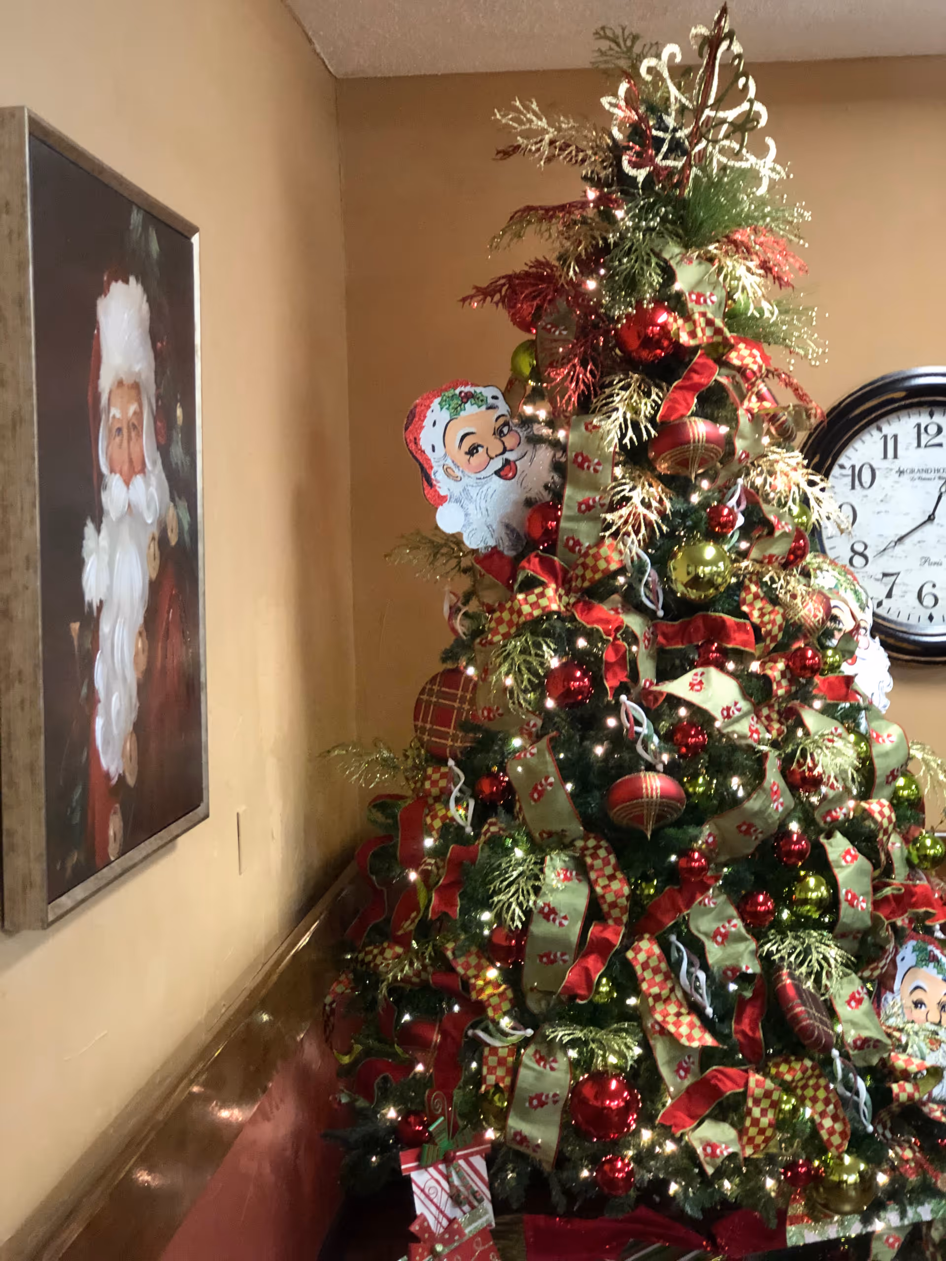 A richly decorated Christmas tree with red and green ribbons and ornaments next to a wall painting of Santa and a large clock in an interior space.