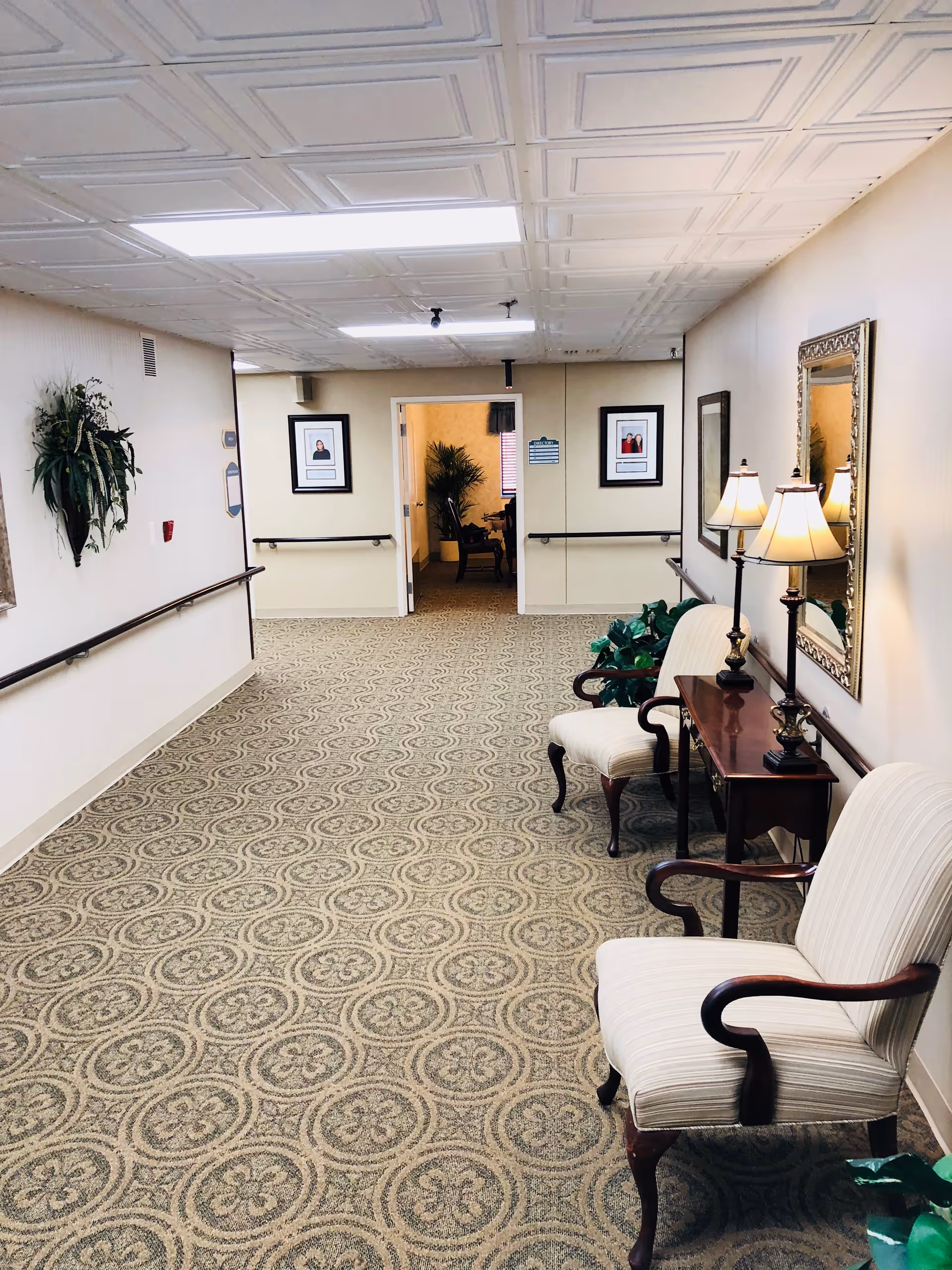 A well-lit hallway in an assisted living facility with patterned carpet, two upholstered armchairs, a wooden side table with two lamps, a large mirror on the wall, framed pictures, and plants. Handrails run along both walls and an open doorway leads to another room with more plants and furniture.