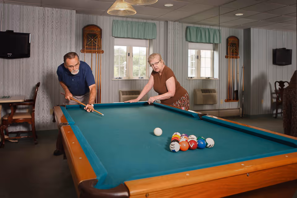 An elderly man and woman playing pool together in a recreational room with two windows, two wall-mounted cue racks, and a television on the wall.