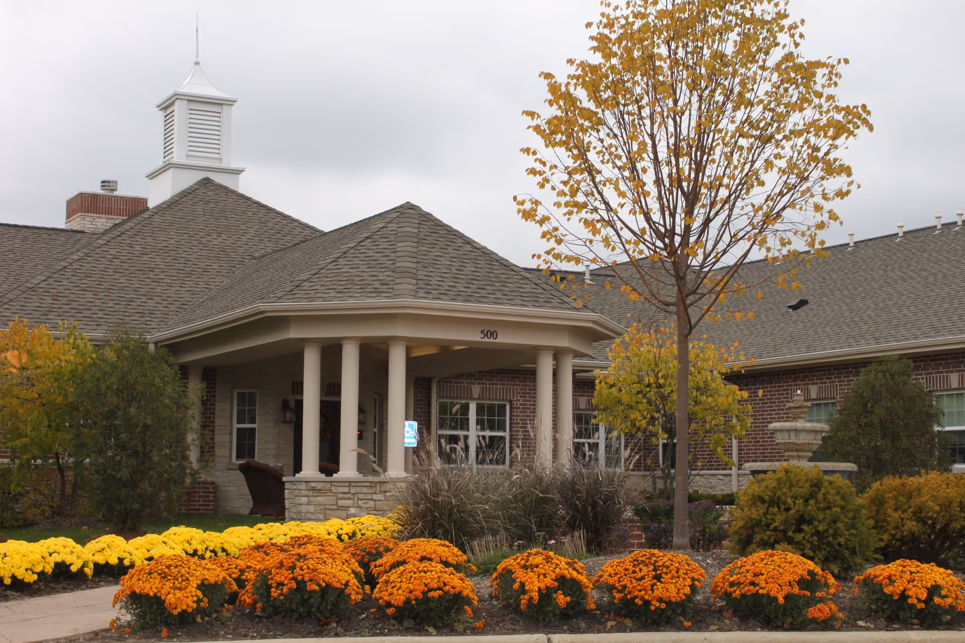 Exterior view of Aspen Woods of Vernon Hills facility showing a brick building with a covered entrance supported by columns, a small tower structure on the roof, and landscaped garden with orange and yellow flowers and trees with autumn foliage.