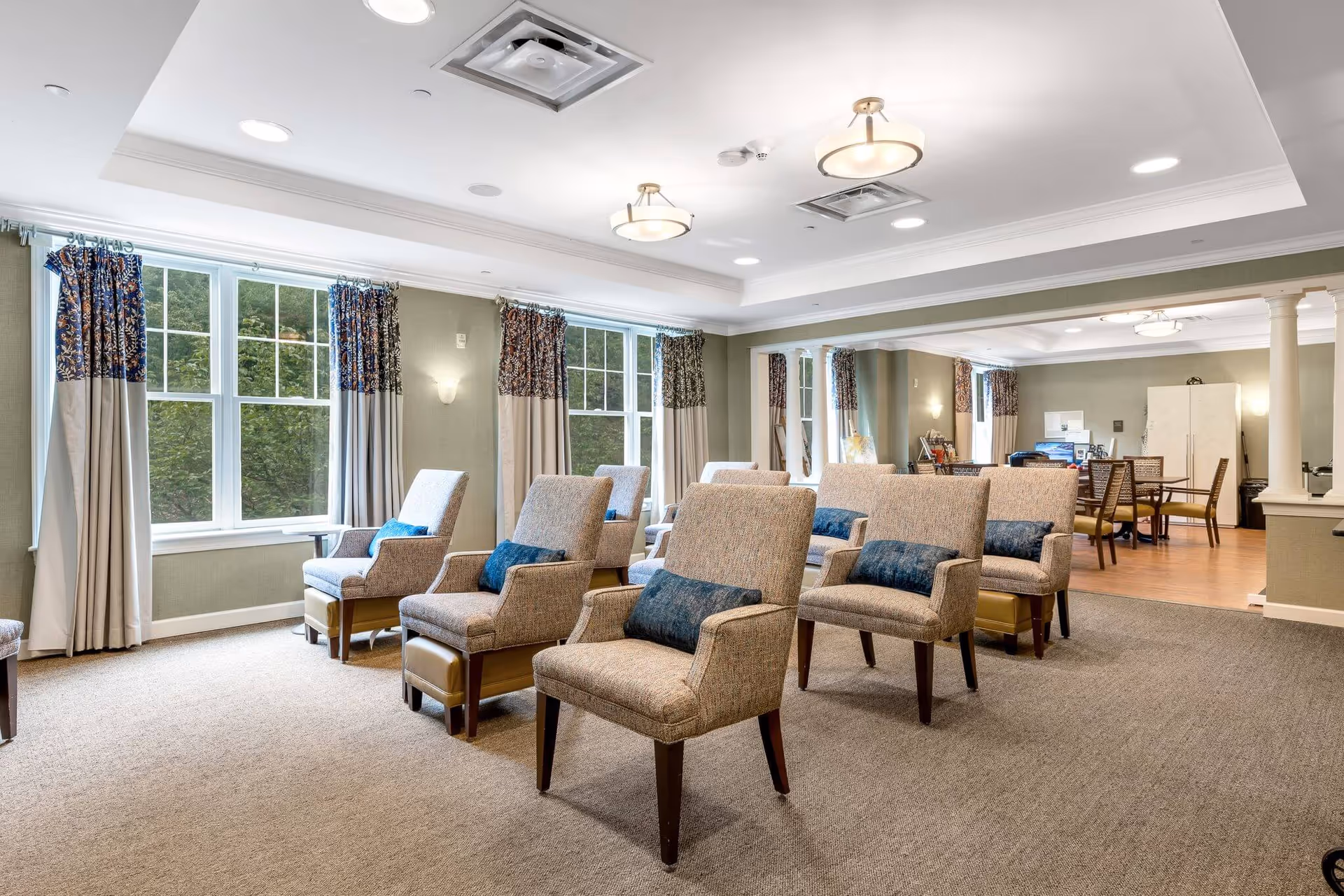 Bright communal lounge with rows of upholstered armchairs and pillows facing a seating area, large windows with curtains, and a dining area in the background.
