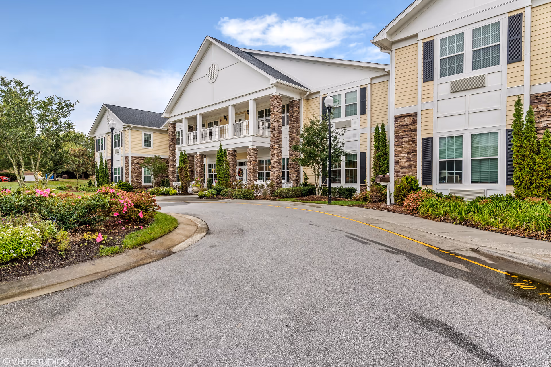 Exterior view of a senior living facility building with beige siding, stone accents, and white trim. The building features a covered entrance with columns and a balcony above. There are landscaped flower beds and shrubs along the driveway leading to the entrance under a partly cloudy sky.