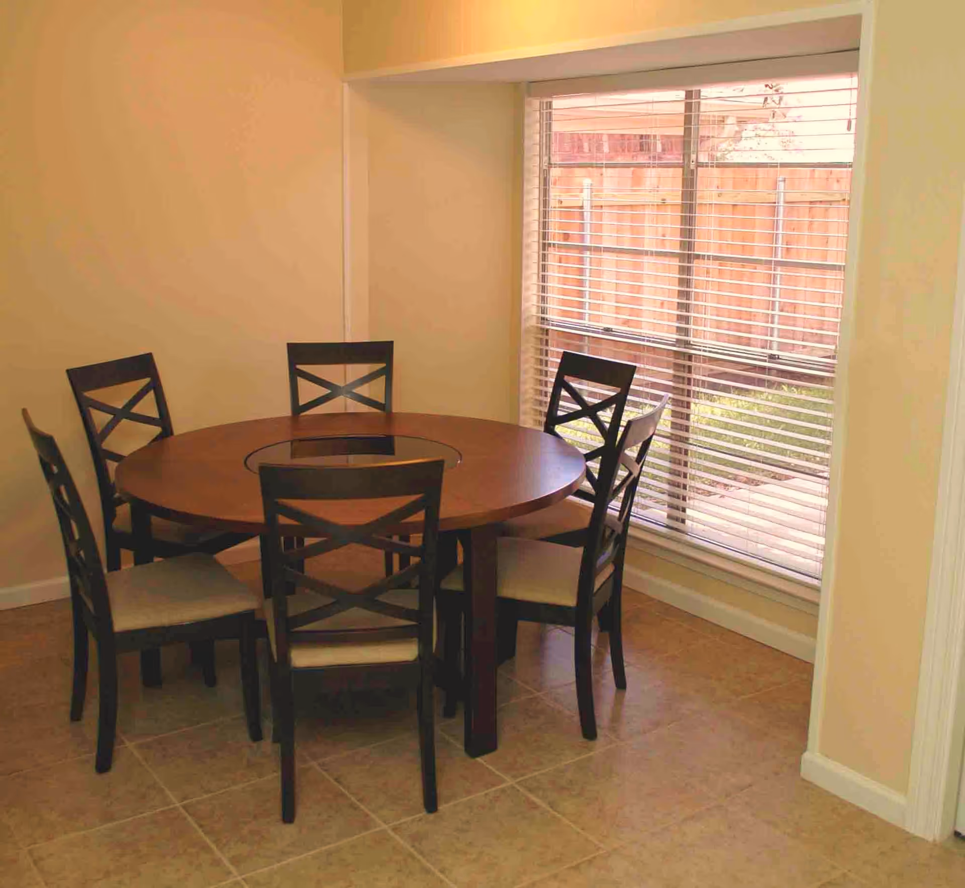 Round wooden dining table with six chairs beside a large window with blinds.