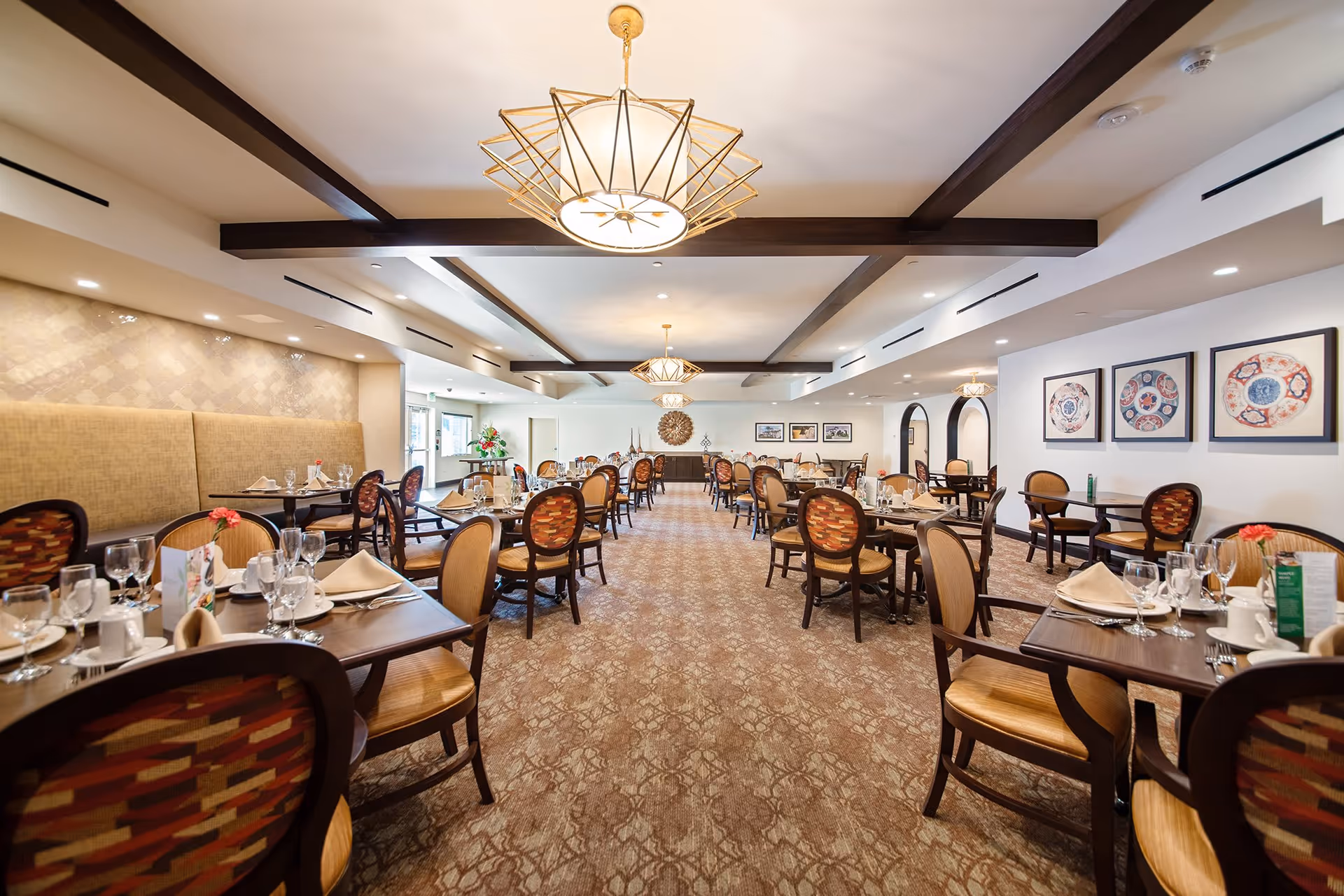 A spacious and elegant dining room with multiple tables set for meals, featuring chairs with patterned upholstery, beige cushioned bench seating along one wall, decorative light fixtures hanging from a ceiling with exposed wooden beams, framed artwork on the walls, and a carpeted floor.