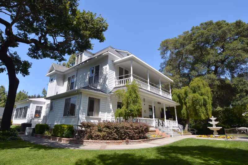 White two-story Victorian-style house with wraparound porches, a manicured lawn, trees, and a fountain in front.