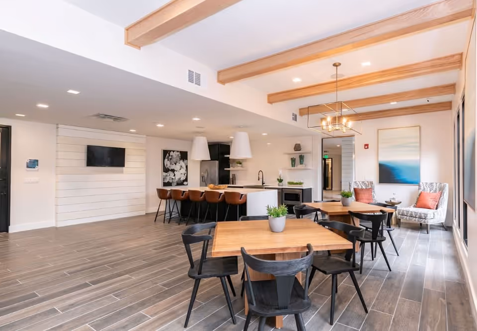 A modern communal dining and kitchen area with wooden tables and black chairs, a kitchen island with brown bar stools, pendant lights, and decorative plants. The room features wooden ceiling beams, a wall-mounted TV, and large windows letting in natural light.