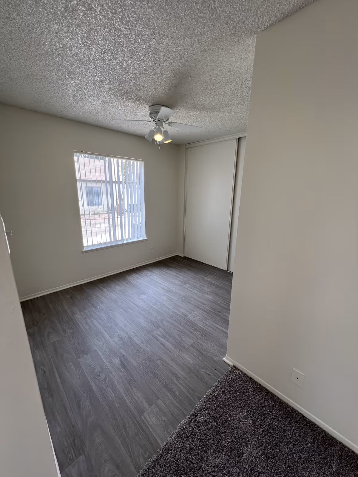 Empty bedroom with a ceiling fan, window with vertical blinds, sliding closet doors, and wood-look flooring.