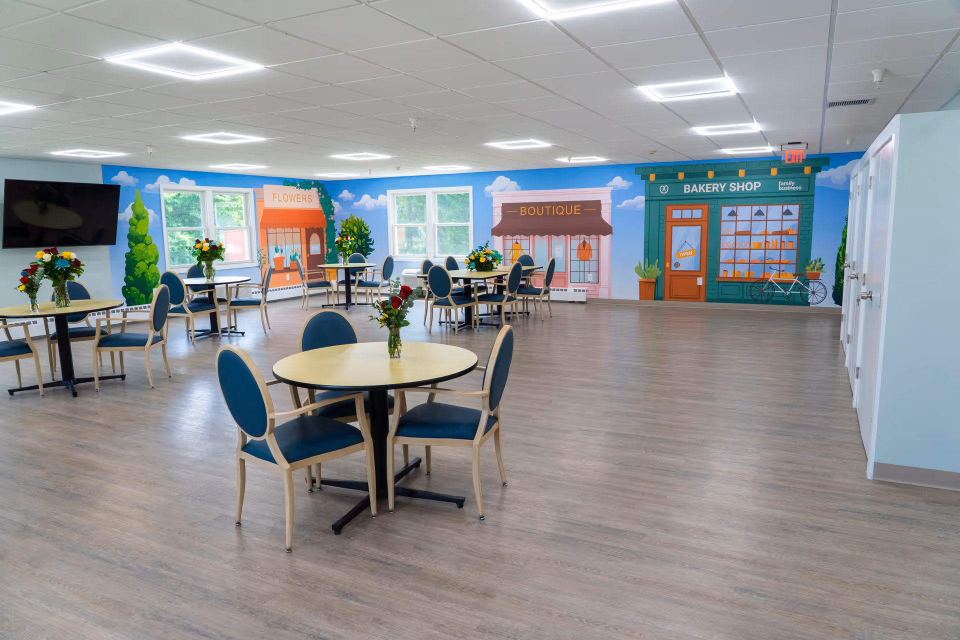 Large bright dining/activity room with round tables and chairs and a colorful mural of storefronts on the far wall.