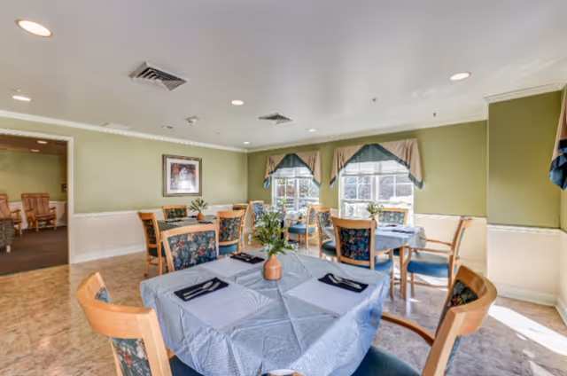 A bright dining room with several tables covered in light blue tablecloths, each set with placemats and napkins. Wooden chairs with floral upholstery surround the tables. The walls are painted light green with white wainscoting, and two windows with valances allow natural light to fill the room. A framed picture hangs on one wall, and a doorway leads to another room with additional seating.