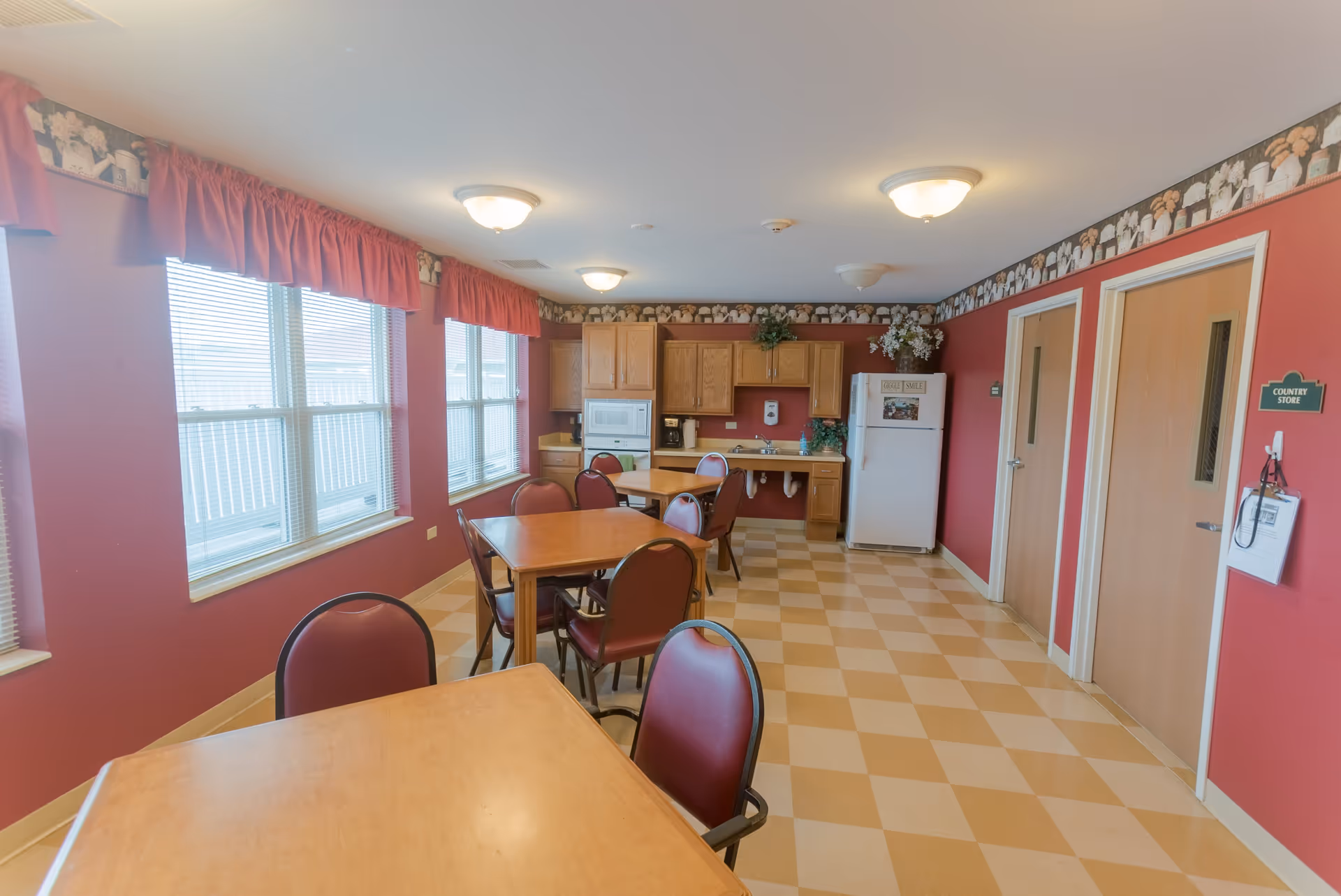 A bright dining area with several wooden tables and red cushioned chairs. The room has large windows with red valance curtains, red walls with floral wallpaper border near the ceiling, and a checkered beige and light brown floor. At the far end, there is a kitchenette with wooden cabinets, a white microwave, a coffee maker, a sink, and a white refrigerator. Two wooden doors are on the right wall, one labeled 'Country Store'.
