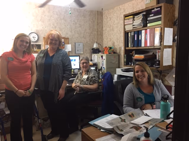 Four women smiling in a small office space with desks, computers, filing cabinets, and shelves filled with binders and papers. The room has beige patterned wallpaper and a ceiling fan.