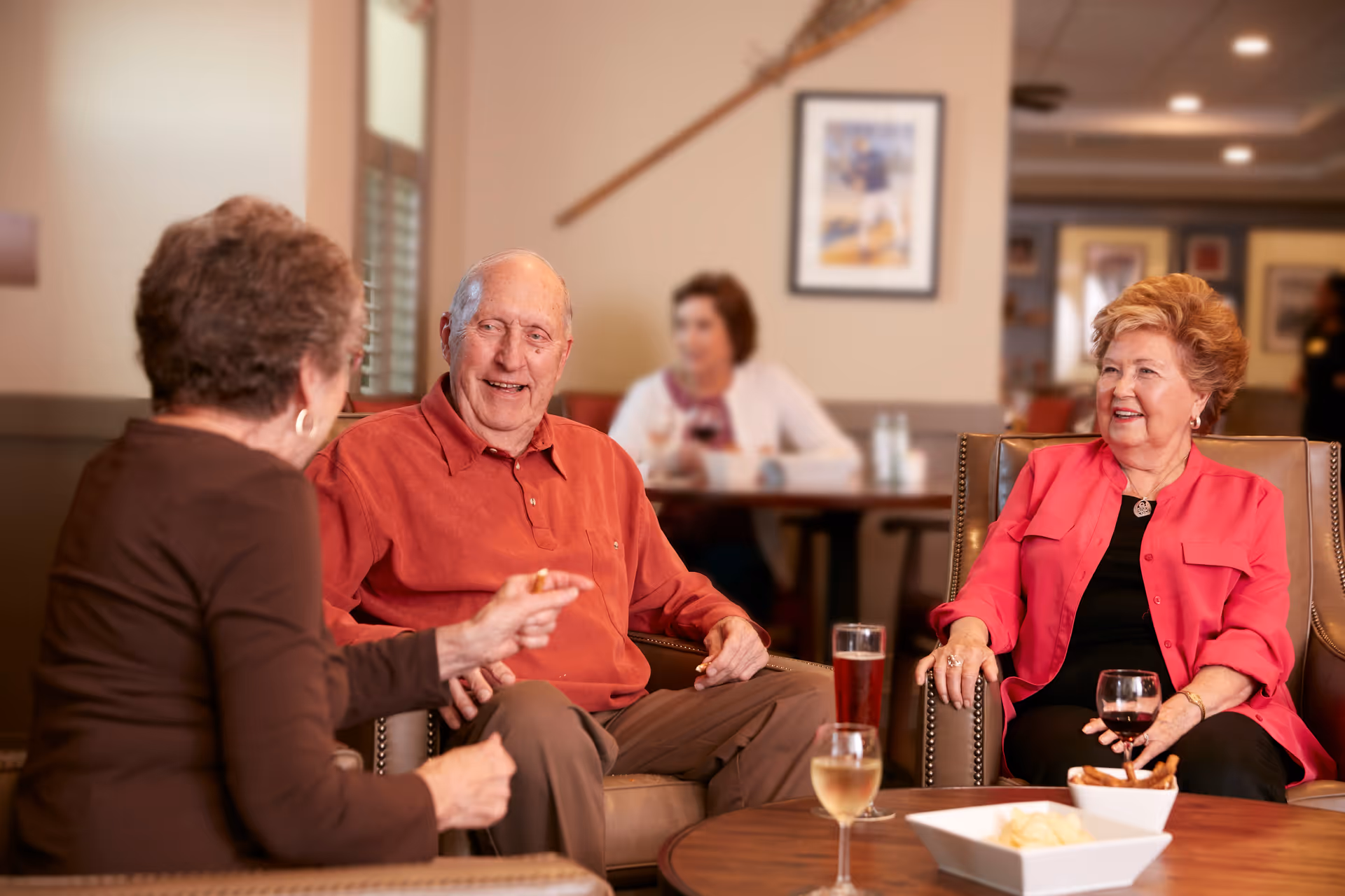 Three elderly people sitting and chatting in a comfortable lounge area. Two women and one man are seated around a wooden table with drinks and snacks. The background shows another person sitting at a table in a softly lit room with framed pictures on the walls.