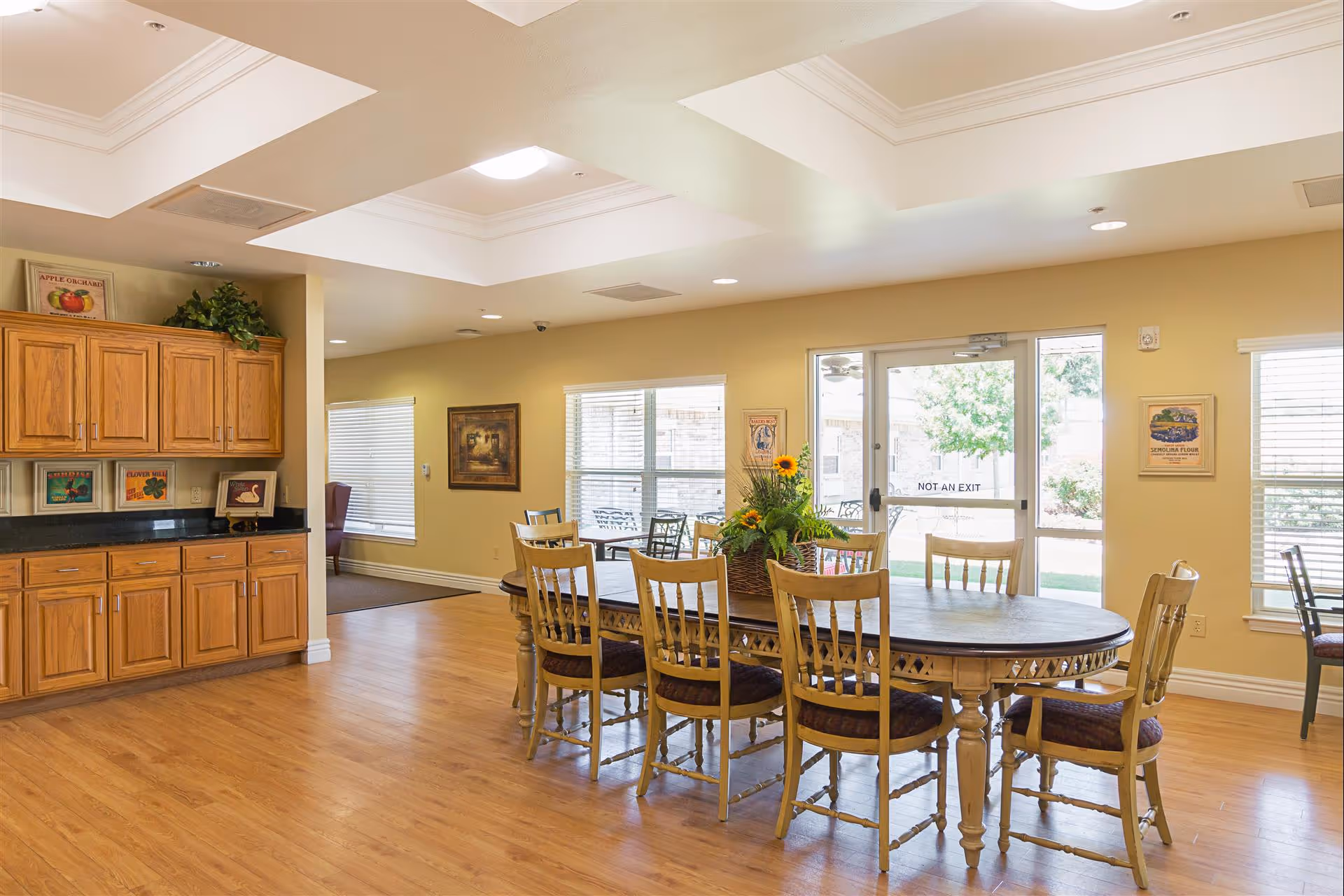 A bright dining room with a large wooden table surrounded by eight chairs. The table has a floral centerpiece with sunflowers. The room has light yellow walls, wooden flooring, and several windows letting in natural light. There are wooden cabinets with framed pictures on the left side and a glass door labeled 'NOT AN EXIT' leading outside.