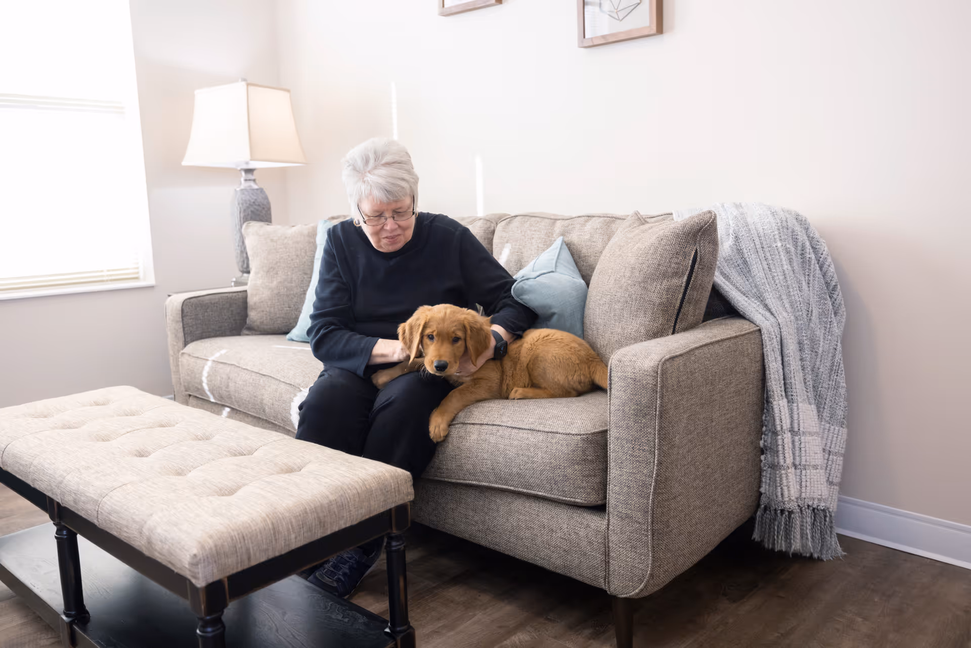 An elderly woman with gray hair sitting on a beige couch in a living room, petting a golden retriever puppy lying beside her. The room has a floor lamp, a window with blinds, a cushioned bench, and a gray throw blanket draped over the couch.