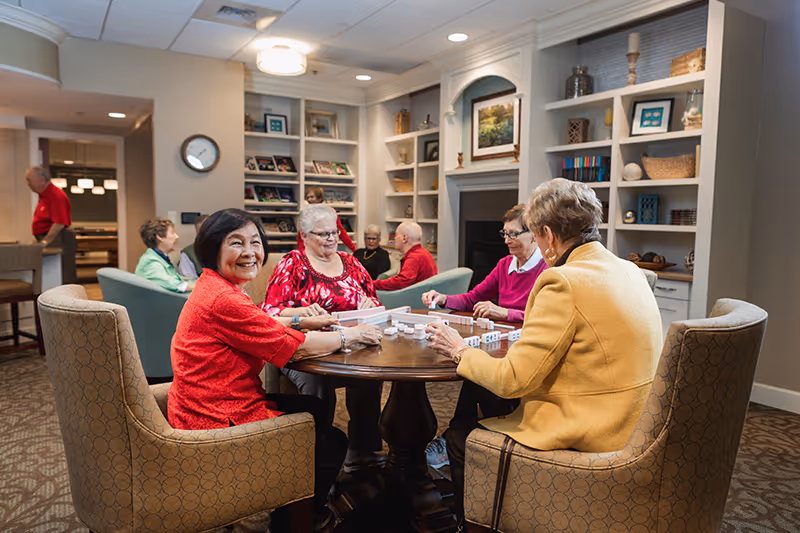 A group of elderly people sitting around a round wooden table playing a game of dominoes in a cozy common area with bookshelves, a fireplace, and comfortable chairs. The room has warm lighting and a welcoming atmosphere.