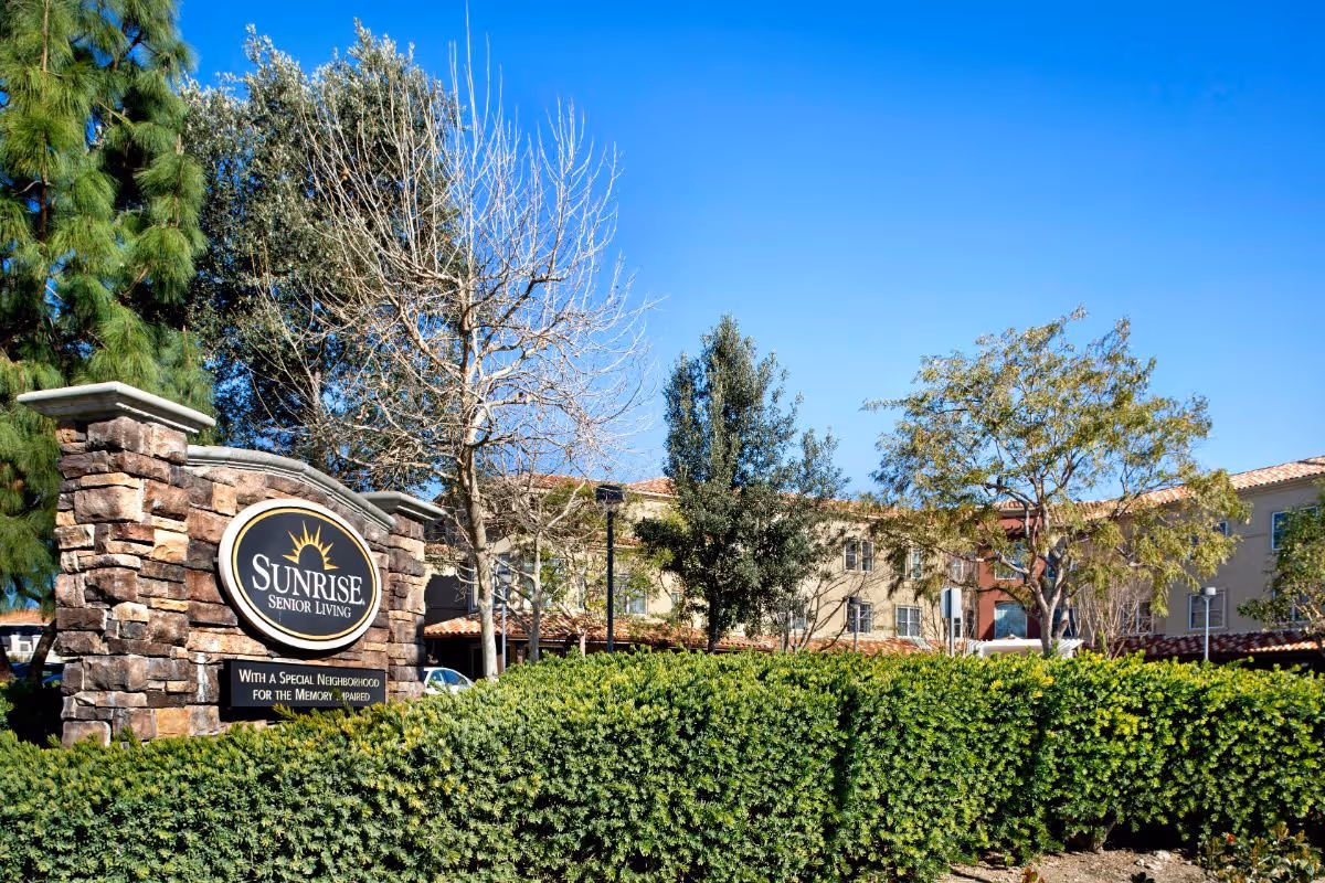 Outdoor view of the entrance sign for Sunrise Senior Living facility, surrounded by green bushes and trees with a clear blue sky in the background. The building is partially visible behind the trees.