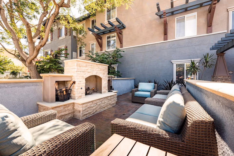 Outdoor patio area at The Vistas at Oxnard Senior Living featuring wicker furniture with light blue cushions arranged around a stone fireplace. The patio is surrounded by a low wall and adjacent to a multi-story building with windows and decorative wooden awnings. Trees and plants add greenery to the space.