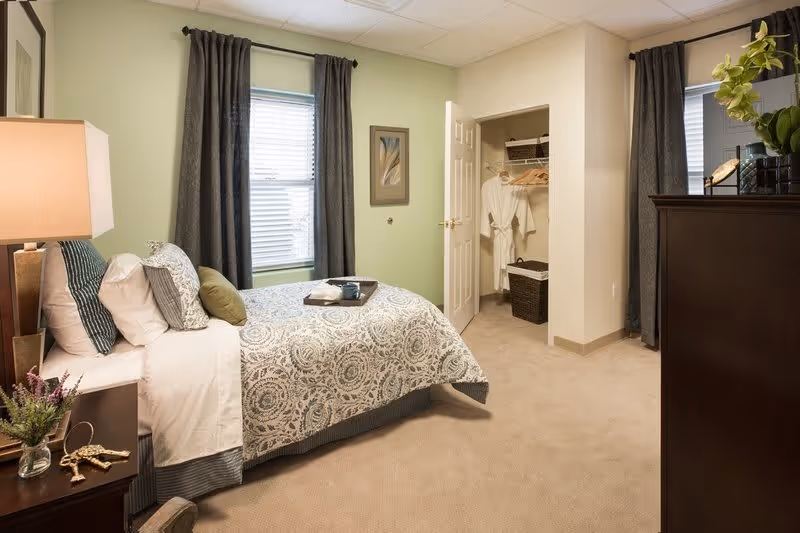 A cozy bedroom with a neatly made bed featuring patterned bedding and multiple pillows. There is a nightstand with a lamp and decorative items next to the bed. The room has two windows with dark curtains, a framed picture on the wall, an open closet with robes and hangers, and a dark wooden dresser with a plant on top.