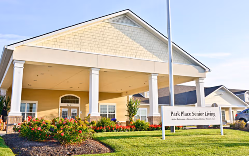 Front entrance of Park Place Senior Living showing a covered portico with columns, landscaped flowerbeds, and a sign on the lawn.