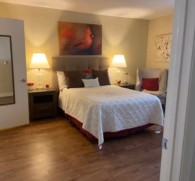 A cozy bedroom with a neatly made bed covered in a white quilt and red bed skirt, flanked by two nightstands each with a lamp. Above the bed hangs a painting of a red cardinal bird. To the right, there is a cushioned armchair with a red pillow and a piece of abstract art on the wall behind it. The floor is wooden, and a mirror is mounted on the left wall near the door.