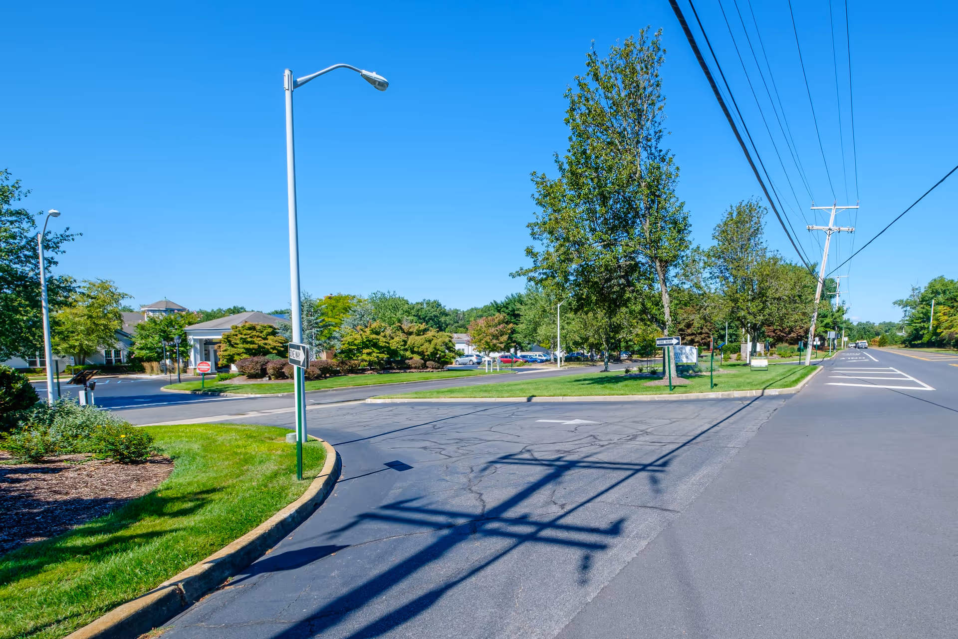 View of the entrance road to Crestwood Manor senior living facility on a clear sunny day, showing a paved driveway, green lawns, trees, street lamps, and a sign for Crestwood Manor near the entrance.